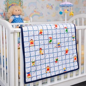 May include: White baby quilt with a grid pattern and colorful embroidered figures. The quilt has a navy blue border and is displayed on a white crib. A doll and mobile are visible in the background, suggesting a nursery setting.