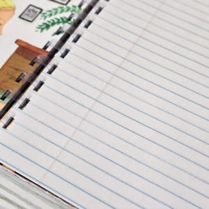 May include: A spiral-bound notebook with white lined paper. The top left corner features a partial illustration of a person with blonde hair, a brown desk, and a green plant. The notebook has a black plastic coil binding along the left edge.
