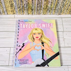 May include: A Little Golden Book Biography of Taylor Swift featuring a blonde illustration of the singer making a heart shape with hands, holding a guitar, and standing near a microphone. The cover text reads Taylor Swift, Wendy Loggia, and Elisa Chavarri.