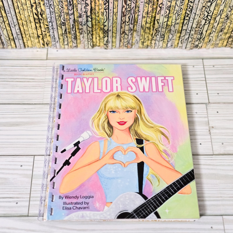 May include: A Little Golden Book Biography of Taylor Swift featuring a blonde illustration of the singer making a heart shape with hands, holding a guitar, and standing near a microphone. The cover text reads Taylor Swift, Wendy Loggia, and Elisa Chavarri.