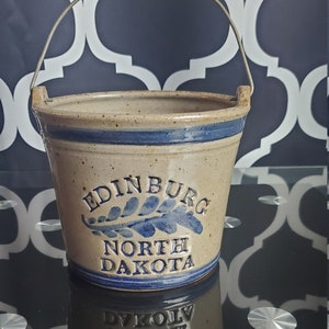 Könnte beinhalten: Ein Steinzeug-Eimer mit Holzgriff und Metallbügeln. Der Eimer ist beige mit blauem Streifen und der Aufschrift "EDINBURG NORTH DAKOTA" mit blauem Blattdesign. Der Eimer spiegelt sich auf einer schwarzen Oberfläche.
