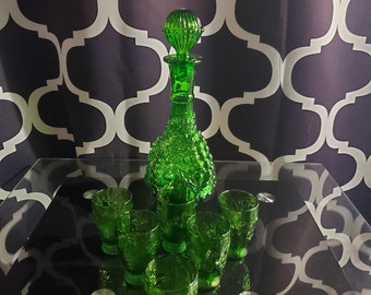 Puede incluir: Un juego de seis vasos de chupito de vidrio verde y una jarra de vidrio verde a juego con un diseño decorativo. La jarra tiene un cuerpo redondo y bulboso y un cuello alto y delgado.
