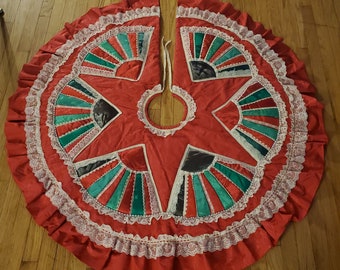 Pode incluir: Uma saia de árvore de Natal vermelha com acabamento em renda branca e desenhos em forma de leque verde e branco.