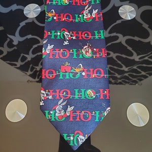 Peut inclure: Une cravate de Noël bleue avec un motif répété de "Ho Ho Ho" et des personnages de dessins animés de Looney Tunes. Les personnages incluent Bugs Bunny, Daffy Duck, Tweety Bird et Sylvester le chat.