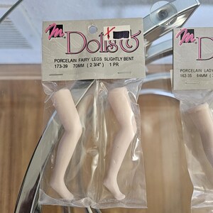 May include: Two pairs of porcelain doll legs, slightly bent, packaged in clear plastic. The packaging reads "Dolls" and "PORCELAIN FAIRY LEGS SLIGHTLY BENT 70MM (2 3/4") 1 PR". The legs are a pale, flesh-toned color.