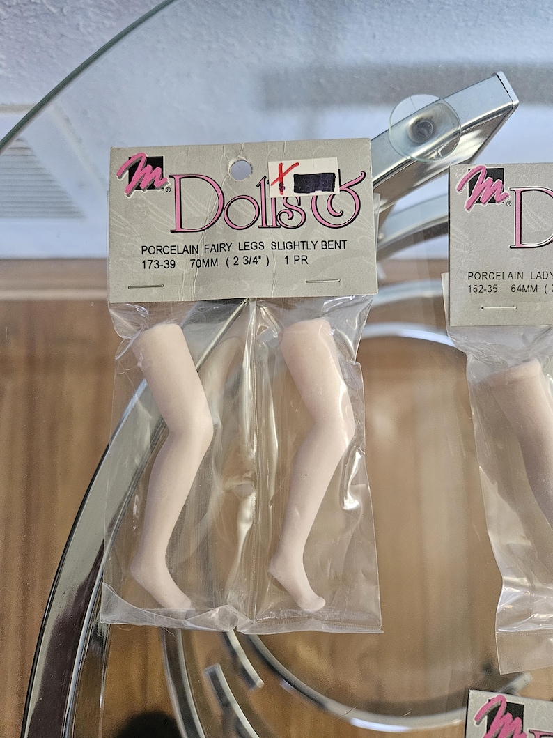 May include: Two pairs of porcelain doll legs, slightly bent, packaged in clear plastic. The packaging reads "Dolls" and "PORCELAIN FAIRY LEGS SLIGHTLY BENT 70MM (2 3/4") 1 PR". The legs are a pale, flesh-toned color.