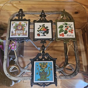 Puede incluir: Cuatro salvamanteles decorativos de azulejos con marcos de metal negro y dorado ornamentados. Los azulejos presentan varios diseños: un reloj, un frutero, frambuesas y un gato. Los salvamanteles se exhiben sobre una mesa de cristal.