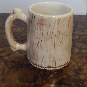 May include: A ceramic coffee mug with a tree trunk design. The mug is light brown with dark brown wood grain details. The handle is shaped like a tree branch.