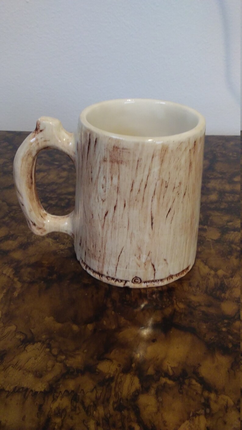 May include: A ceramic coffee mug with a tree trunk design. The mug is light brown with dark brown wood grain details. The handle is shaped like a tree branch.