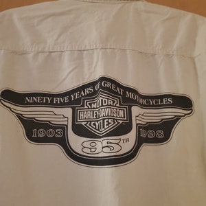 Op de afbeelding: Een wit overhemd met lange mouwen en knopen met een zwart Harley-Davidson-logo op de rug. Het logo bevat de woorden "Ninety Five Years of Great Motorcycles", "Harley-Davidson" en "1903-1998".