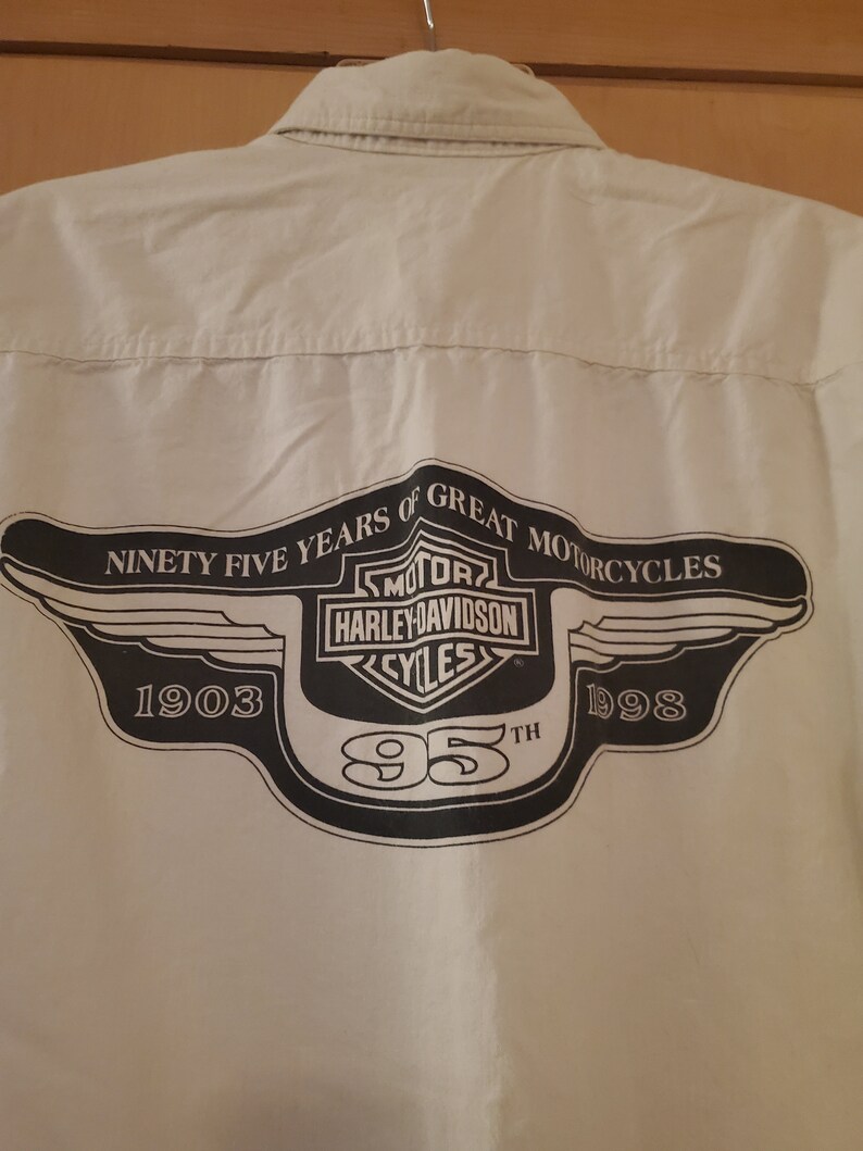 Op de afbeelding: Een wit overhemd met lange mouwen en knopen met een zwart Harley-Davidson-logo op de rug. Het logo bevat de woorden "Ninety Five Years of Great Motorcycles", "Harley-Davidson" en "1903-1998".