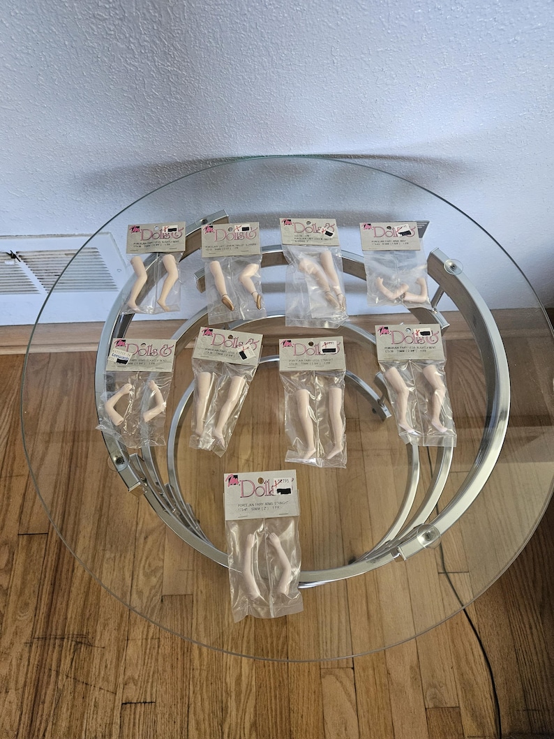 May include: A collection of doll limbs, including arms and legs, packaged in clear plastic. The limbs are a pale flesh tone and are displayed on a round glass table with a silver metal base. Each package has a label with the word "Dolls" on it.