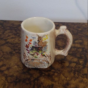 May include: A white ceramic mug with a tree trunk design and a brown handle. The mug features a colorful illustration of a deer, birds, and squirrels in a forest setting. The text "The Flags" is printed on the bottom of the mug.