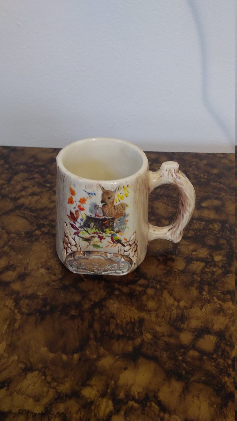 May include: A white ceramic mug with a tree trunk design and a brown handle. The mug features a colorful illustration of a deer, birds, and squirrels in a forest setting. The text "The Flags" is printed on the bottom of the mug.
