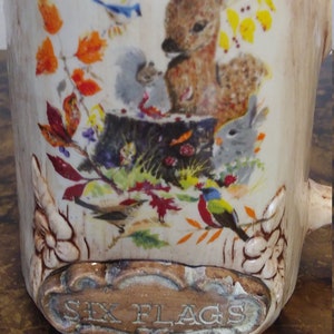 May include: A ceramic mug with a woodland scene featuring a deer, a squirrel, a rabbit, and birds. The mug has a brown and white glaze and the words "Six Flags" are printed on the bottom.