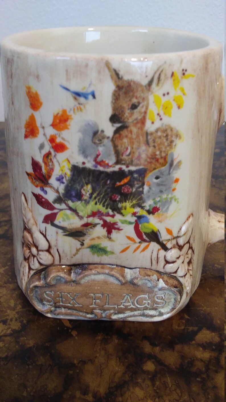 May include: A ceramic mug with a woodland scene featuring a deer, a squirrel, a rabbit, and birds. The mug has a brown and white glaze and the words "Six Flags" are printed on the bottom.