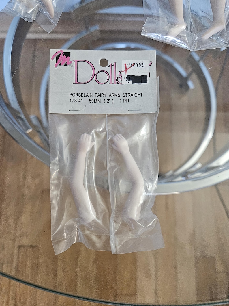 May include: A package of porcelain doll arms, straight, in a clear plastic bag. The label reads "Dolls" and "Porcelain Fairy Arms Straight 173-41 50mm (2") 1 PR". The arms are white and appear to be for a fairy doll.