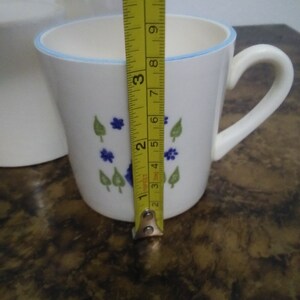Może przedstawiać: Biały kubek ceramiczny z niebiesko-zielonym wzorem kwiatowym. Kubek ma uchwyt i niebieski rant. Kubek ma 10 cm wysokości.