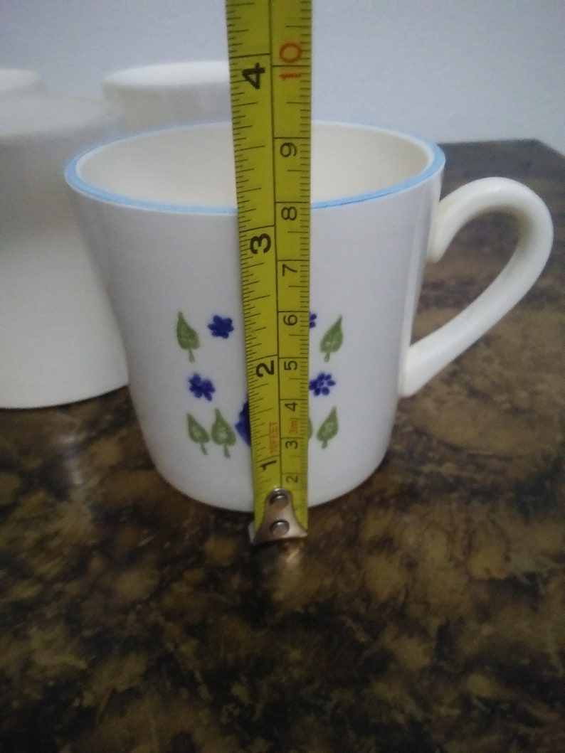 Może przedstawiać: Biały kubek ceramiczny z niebiesko-zielonym wzorem kwiatowym. Kubek ma uchwyt i niebieski rant. Kubek ma 10 cm wysokości.