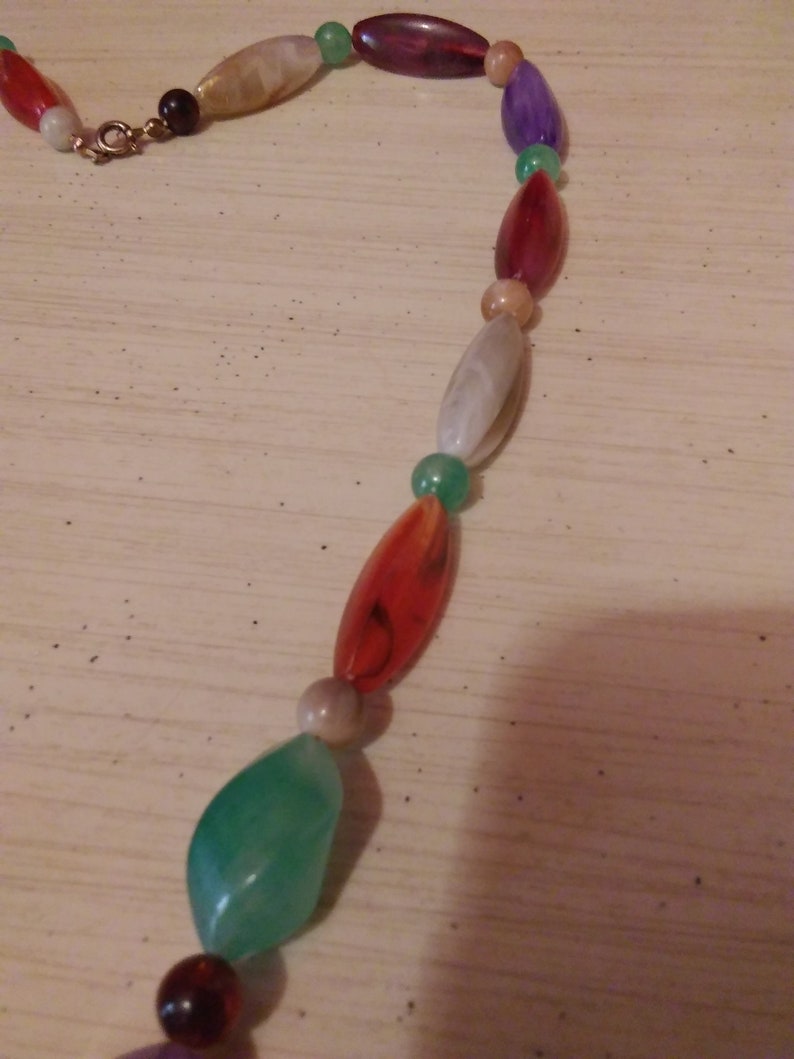 Pu&ograve; includere: Una collana di perline multicolori con perline di forma ovale e a goccia. Le perline sono in vetro e hanno una variet&agrave; di colori, tra cui rosso, verde, viola e bianco.