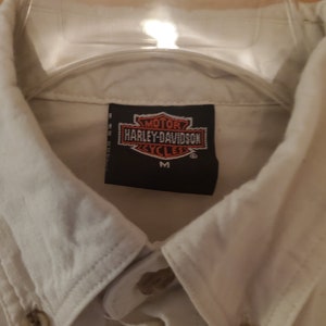 Op de afbeelding: Wit overhemd met knopen en een Harley-Davidson logo patch op de borst. De patch is zwart met witte letters en een rood en oranje logo. Het shirt is maat M.