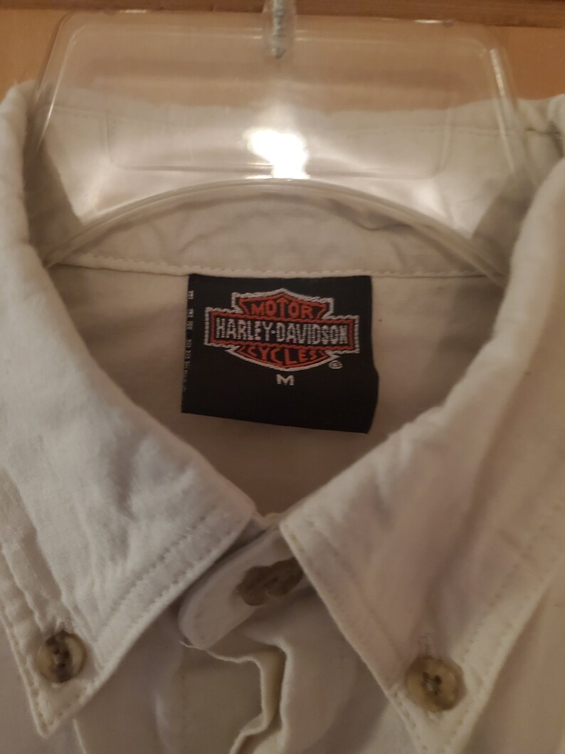 Op de afbeelding: Wit overhemd met knopen en een Harley-Davidson logo patch op de borst. De patch is zwart met witte letters en een rood en oranje logo. Het shirt is maat M.