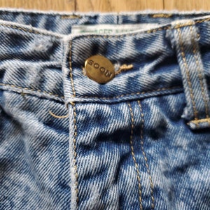May include: Close-up of a pair of light blue denim jeans with a gold button that says "SOON" on it.