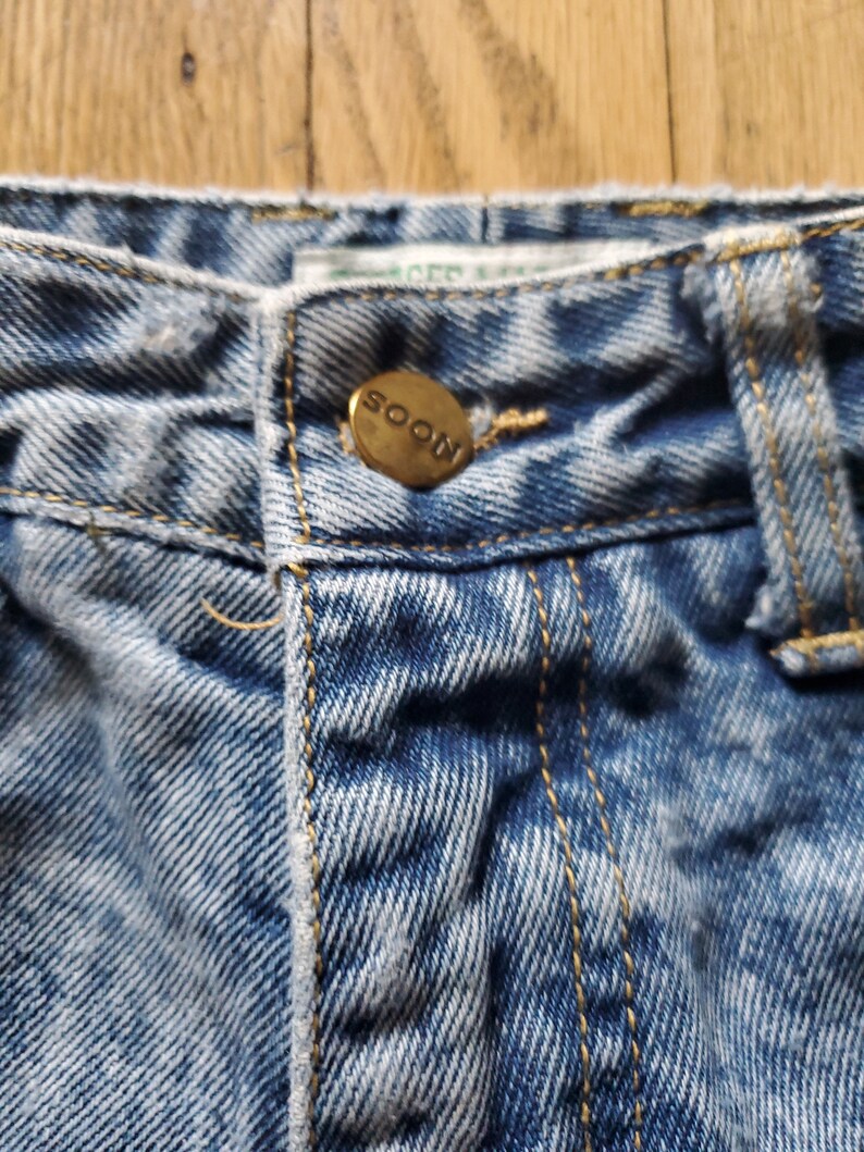 May include: Close-up of a pair of light blue denim jeans with a gold button that says "SOON" on it.