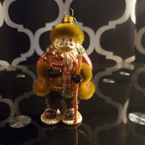 Puede incluir: Un adorno de Navidad de vidrio que representa a Papá Noel con un abrigo rojo y verde, barba blanca y ribete de piel. Lleva un bastón y un pequeño osito de peluche. El adorno tiene un acabado de purpurina dorada.