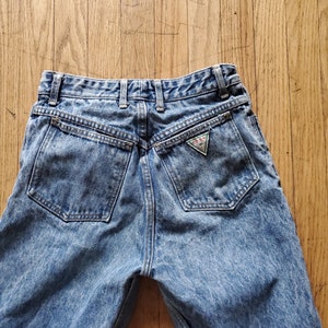 May include: A pair of light blue denim jeans with a faded wash and a triangular Guess logo patch on the back pocket.