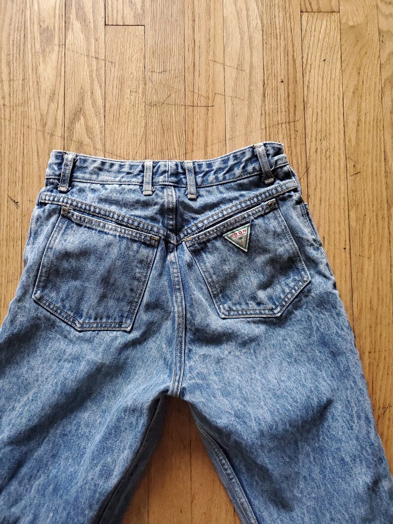 May include: A pair of light blue denim jeans with a faded wash and a triangular Guess logo patch on the back pocket.