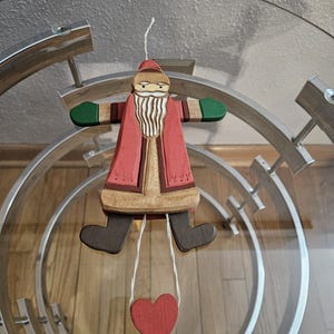 May include: Wooden Santa Claus ornament with a red and white suit, brown beard, and green mittens. The ornament is hanging from a string with a red heart at the bottom.