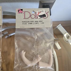 May include: A package containing two porcelain fairy arms, bent at the elbow. The arms are a pale flesh tone and are 1 5/8 inches (41mm) long. The package has a label that reads "Doll's" and includes the product description "Porcelain Fairy Arms Bent".