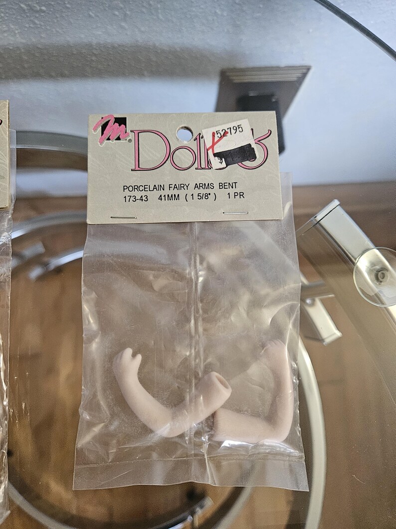 May include: A package containing two porcelain fairy arms, bent at the elbow. The arms are a pale flesh tone and are 1 5/8 inches (41mm) long. The package has a label that reads "Doll's" and includes the product description "Porcelain Fairy Arms Bent".
