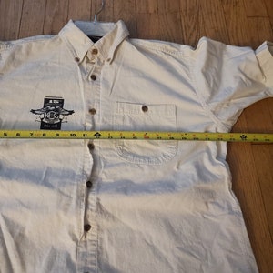 Op de afbeelding: Een wit overhemd met lange mouwen en knopen, met een zakje op de linkerborst. Het shirt heeft een logo op de linkerborst met de tekst "15th Anniversary 1983-1998 K.C.G."