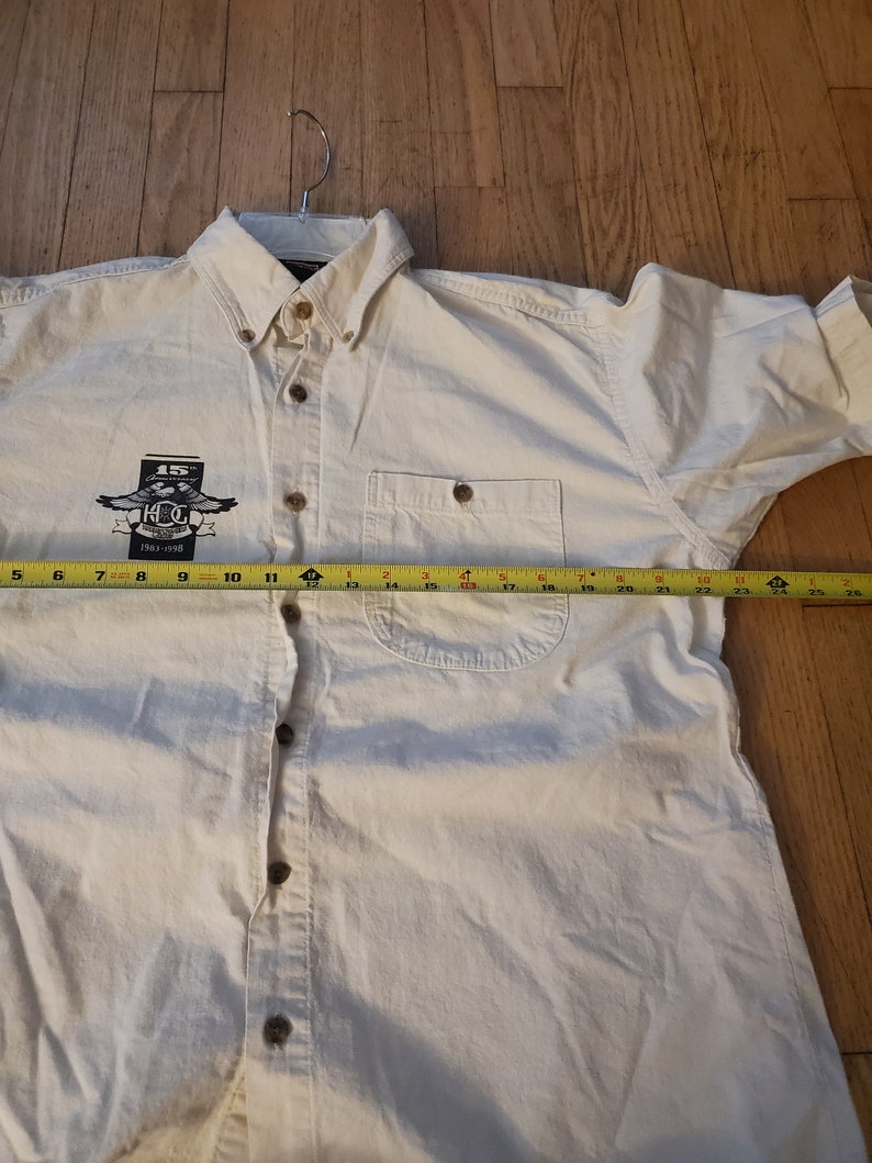 Op de afbeelding: Een wit overhemd met lange mouwen en knopen, met een zakje op de linkerborst. Het shirt heeft een logo op de linkerborst met de tekst "15th Anniversary 1983-1998 K.C.G."