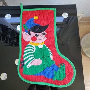 May include: A red and green quilted Christmas stocking with a cartoon elf wearing a green hat and holding a green plant. The stocking has a white and green striped shirt and blue pants. The text "WELLINGTON" is visible on the stocking.