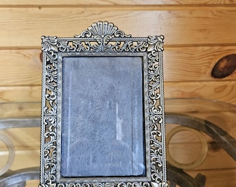 May include: Ornate, antique-style picture frame with a silver finish. The frame features intricate scrollwork and a decorative top. The frame is empty, with a textured, gray backing visible through the glass. A classic piece for displaying photos or artwork.