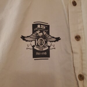 Op de afbeelding: Een wit overhemd met knopen en een zwart-wit logo met de tekst "15th Anniversary" en "Harley Owners Group" met een adelaarontwerp en de jaren "1983-1998".