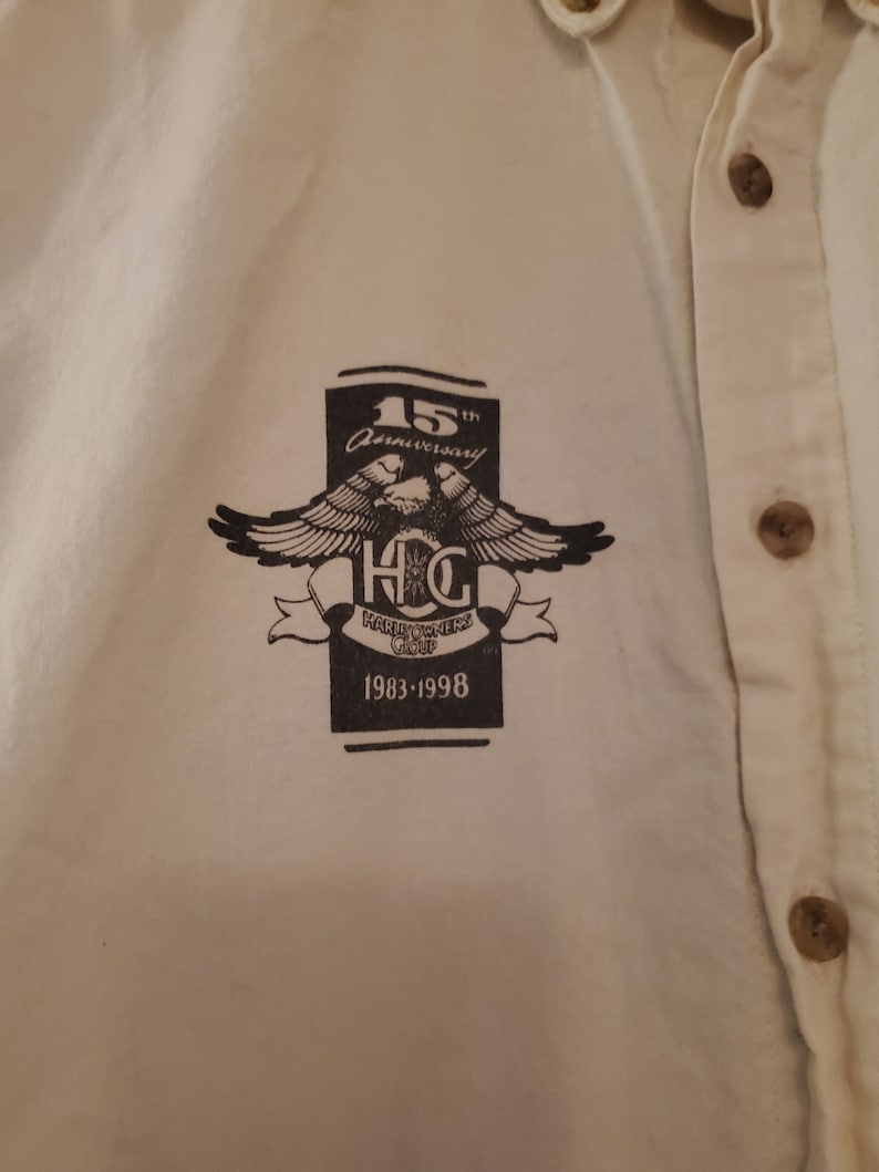 Op de afbeelding: Een wit overhemd met knopen en een zwart-wit logo met de tekst "15th Anniversary" en "Harley Owners Group" met een adelaarontwerp en de jaren "1983-1998".