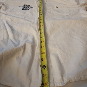 Op de afbeelding: Een wit overhemd met lange mouwen en knopen, met een zak aan de linkerkant. Het shirt heeft een afbeelding van een blikje met de tekst "The Real Thing". Een meetlint is langs het midden van het shirt geplaatst en meet 68,58 cm.