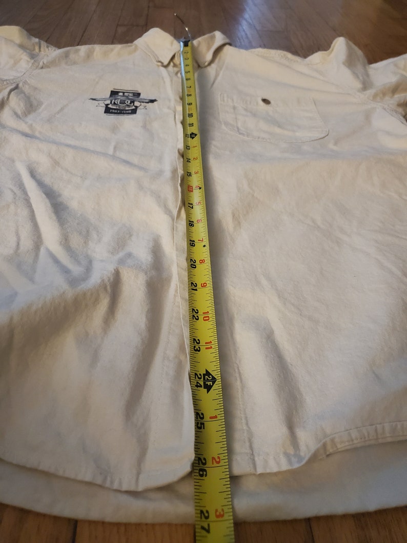 Op de afbeelding: Een wit overhemd met lange mouwen en knopen, met een zak aan de linkerkant. Het shirt heeft een afbeelding van een blikje met de tekst "The Real Thing". Een meetlint is langs het midden van het shirt geplaatst en meet 68,58 cm.