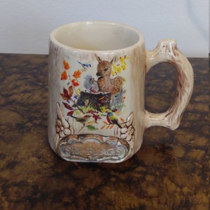 May include: A ceramic mug with a tree trunk design and a handle shaped like a branch. The mug features a colorful illustration of a deer, birds, and other woodland creatures. The text "Six Flags" is printed on the bottom of the mug.