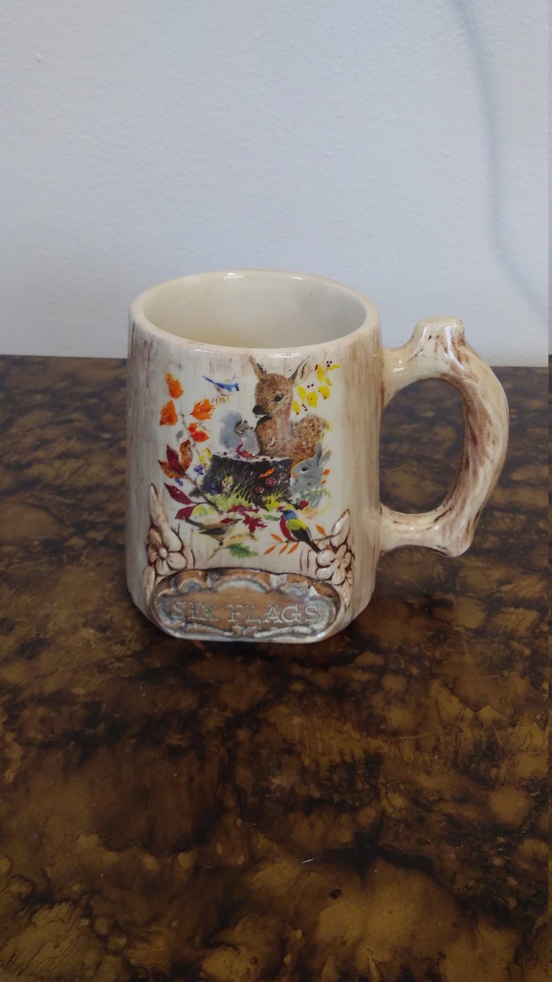 May include: A ceramic mug with a tree trunk design and a handle shaped like a branch. The mug features a colorful illustration of a deer, birds, and other woodland creatures. The text "Six Flags" is printed on the bottom of the mug.