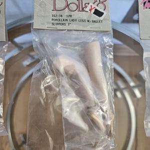 May include: A package containing porcelain lady legs with ballet slippers. The package has a label that reads "Dolls &" and "PORCELAIN LADY LEGS W/ BALLET SLIPPERS 3". The legs are a light skin tone, and the ballet slippers are gold.