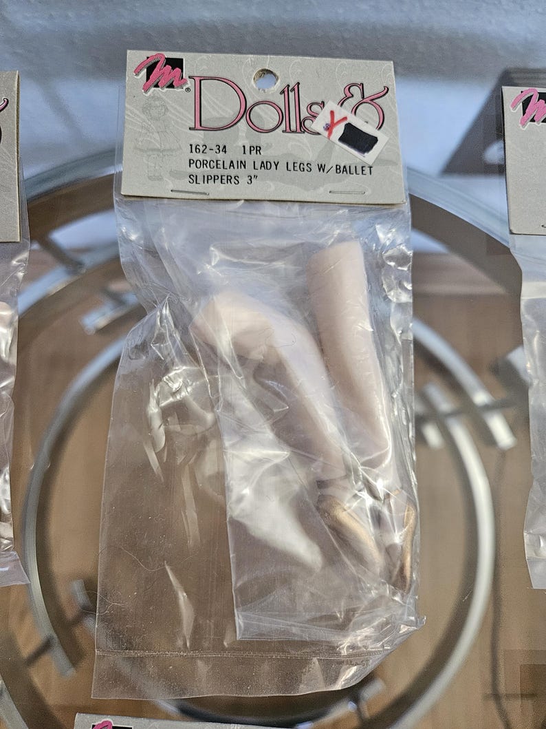 May include: A package containing porcelain lady legs with ballet slippers. The package has a label that reads "Dolls &" and "PORCELAIN LADY LEGS W/ BALLET SLIPPERS 3". The legs are a light skin tone, and the ballet slippers are gold.