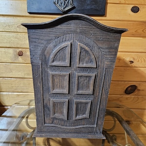 May include: A dark brown wooden cabinet with a decorative arched top and a window-like design on the front. The window is divided into six panels. The cabinet is placed on a glass table.