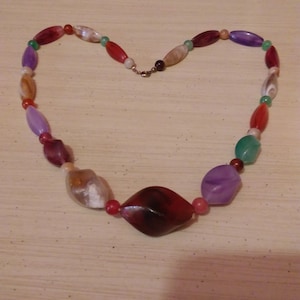 Pu&ograve; includere: Una collana di perline multicolori con una grande perla sfaccettata di colore rosso scuro al centro. La collana &egrave; realizzata con perline di varie tonalit&agrave; di rosso, verde, viola e bianco.