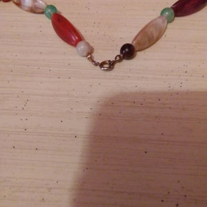 Pu&ograve; includere: Primo piano di una collana con perline ovali rosse, bianche, verdi e marroni. La collana ha una chiusura dorata.