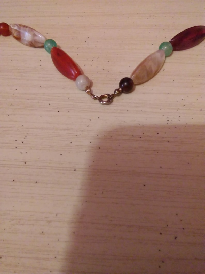 Pu&ograve; includere: Primo piano di una collana con perline ovali rosse, bianche, verdi e marroni. La collana ha una chiusura dorata.
